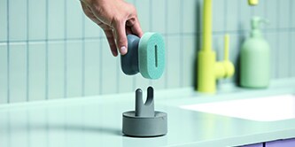 Hand placing a Brite by Scotch-Brite™ Palm Scrubber onto a sponge stand on the kitchen countertop beside the sink.
